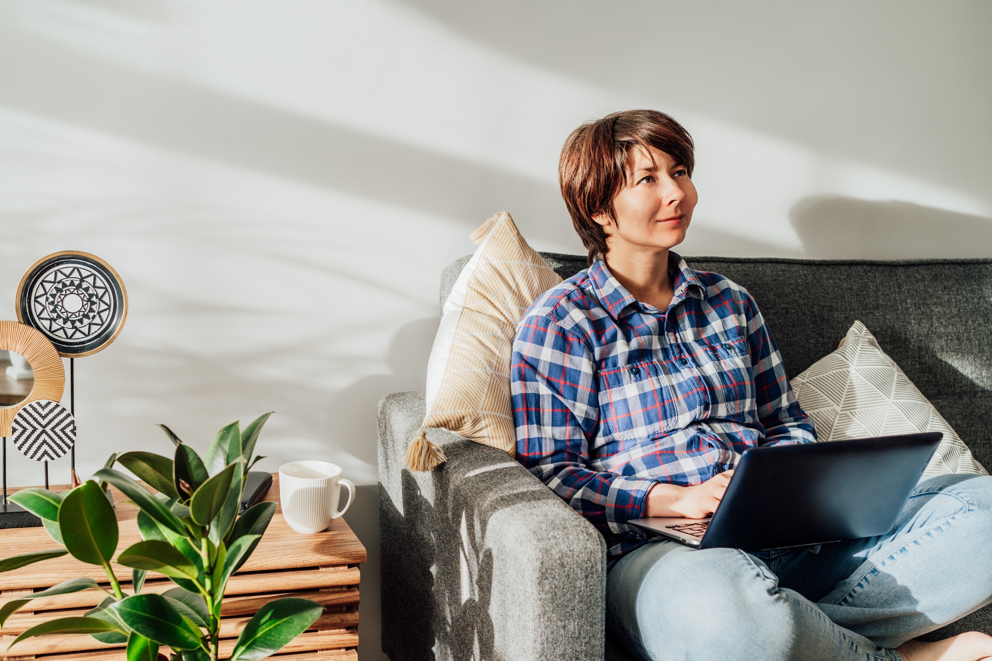 Deep in thoughts woman using laptop at home sitting on sofa, couch in sunlight. Idea Brainstorm.